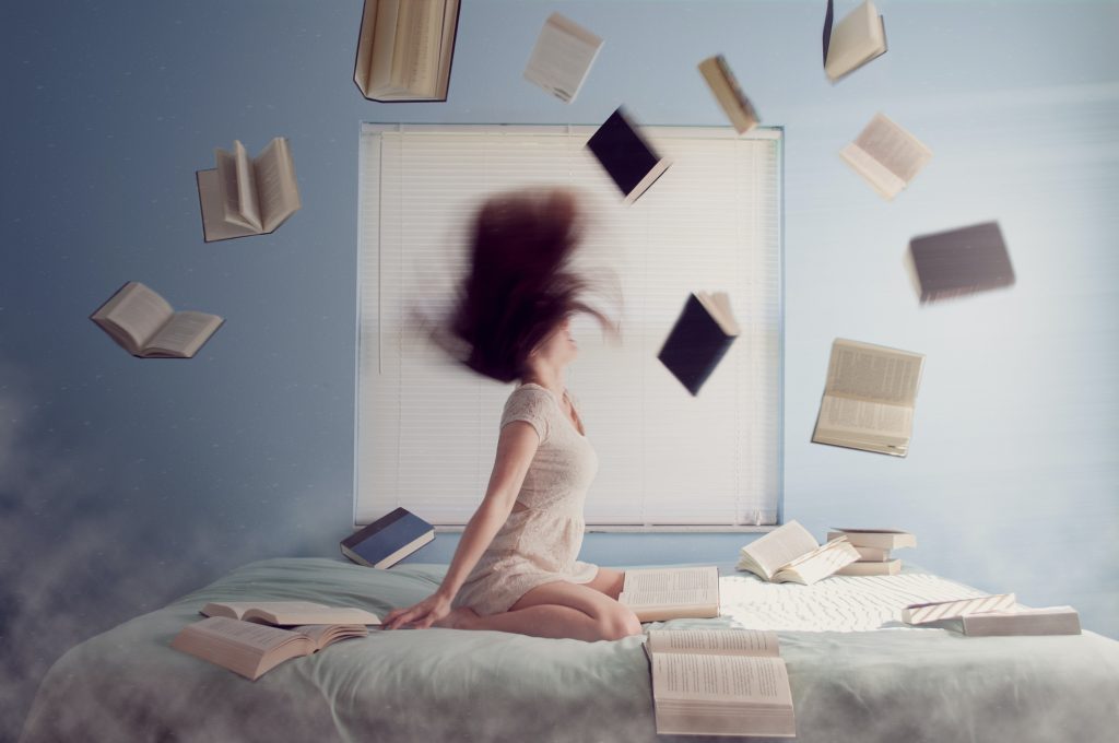 Frau sitzt auf Bett mit etlichen Büchern, welche um sie herumfliegen